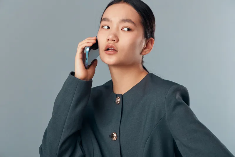 Young woman in business attire talking on smartphone, looking away with focused expression on gray background. Female professional using mobile device for communication and work.