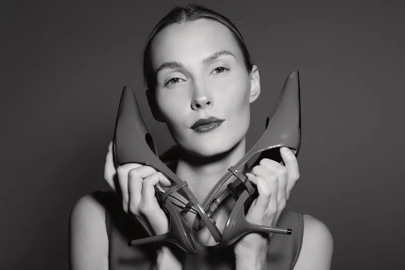 Serious white woman with pale skin holds high heel shoes close to face, posing in studio. Concept for fashion, elegance, and footwear style in black and white photography.