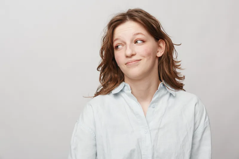 Young woman with wavy brown hair wearing white shirt looks sideways with slight smile on light grey background. Concept of casual style, emotion, and natural beauty portrait.