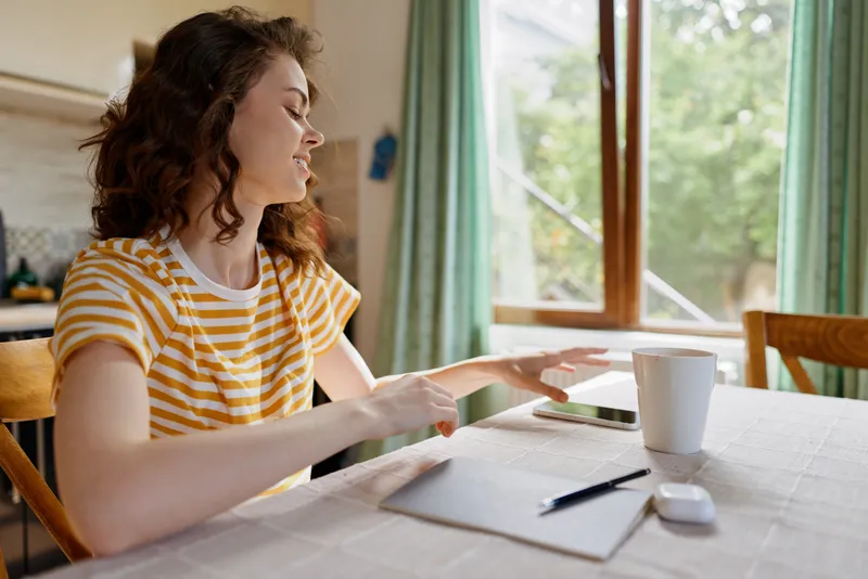 Cheerful woman at home, seated at a sunlit table, reaching forward with relaxed posture, striped shirt, coffee mug nearby, natural colors, cozy lifestyle moment