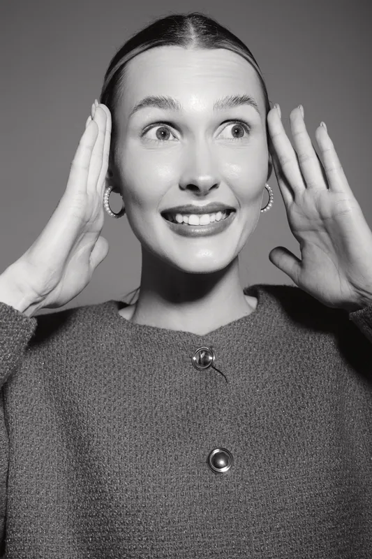 Young white woman with light skin and earrings smiles widely and holds hands near her face. Black and white portrait captures joyful positive emotion and expression.