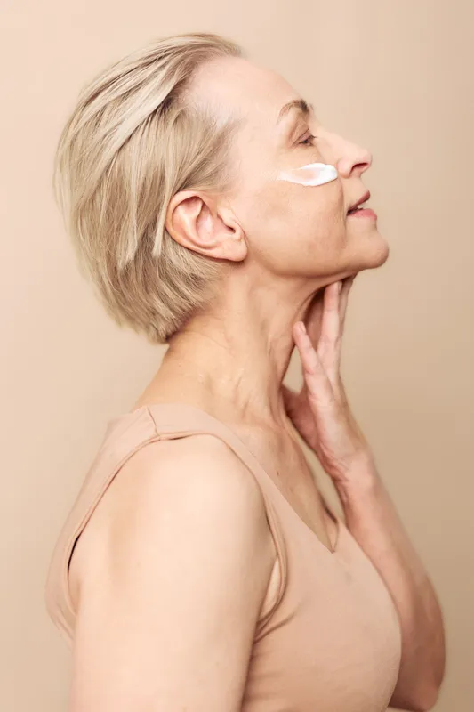 Mature woman with short blonde hair applies face cream on cheek, touching neck gently. Soft beige background creates calm mood. Concept of skincare, aging beauty, wellness and self-care.