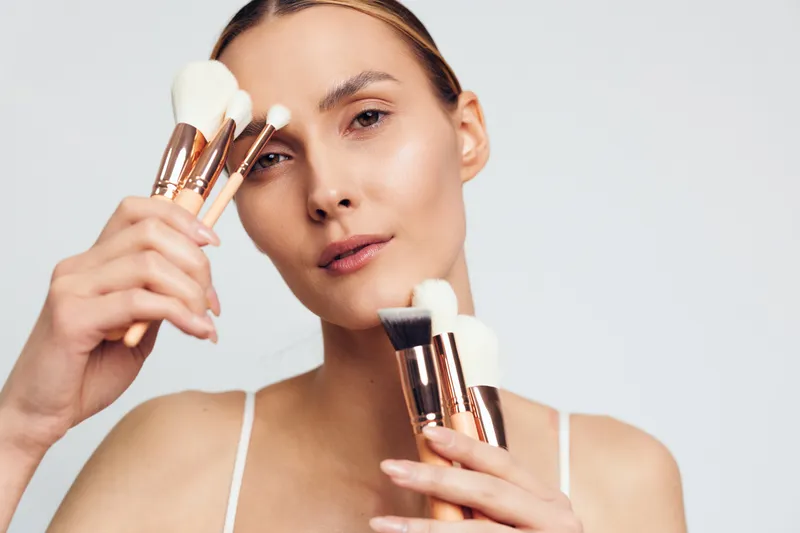 Young woman holding multiple makeup brushes close to her face, neutral expression, soft lighting, clean background, beauty tools, skincare routine concept.
