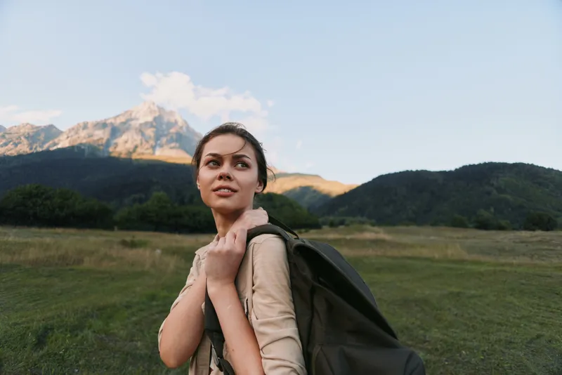 Young woman with a backpack stands in an expansive field as distant mountains rise under a clear sky, conveying adventure, curiosity, and a sense of exploration.