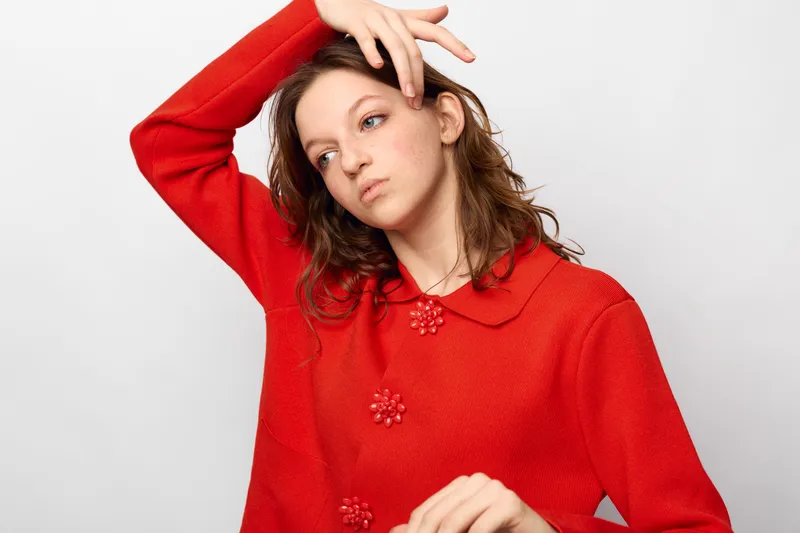 Young woman in red sweater poses with hand on forehead, looking away thoughtfully. Studio portrait on white background. Concept of fashion, emotion, and casual style.