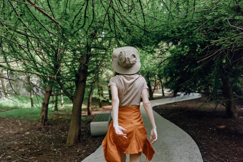 Woman walking forest path with hat and skirt, back view on a curved trail surrounded by trees and greenery for peaceful outdoor leisure and gentle exploration.
