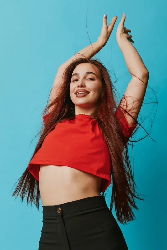 Young woman with long hair wearing red crop top and black pants posing with raised arms and closed eyes against blue background for fashion and lifestyle portrait.