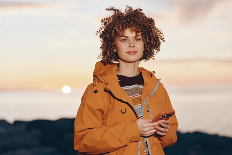 Woman lifestyle portrait outdoors at sunset, curly hair, orange jacket, holding smartphone, looking at camera, scenic mountains in distance, warm evening light, casual travel mood, today