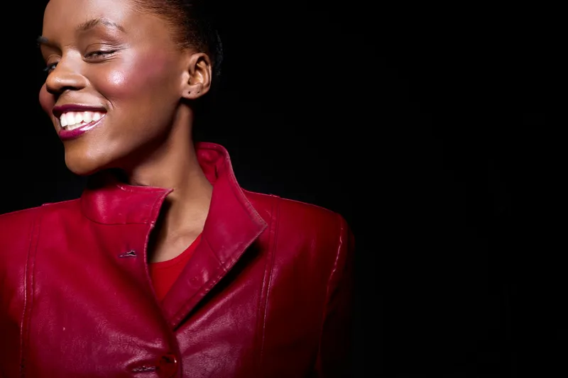 Portrait of a smiling dark skinned African woman with shiny skin wearing a red leather jacket. Concept of beauty, fashion, confidence, and positive emotion on black background.