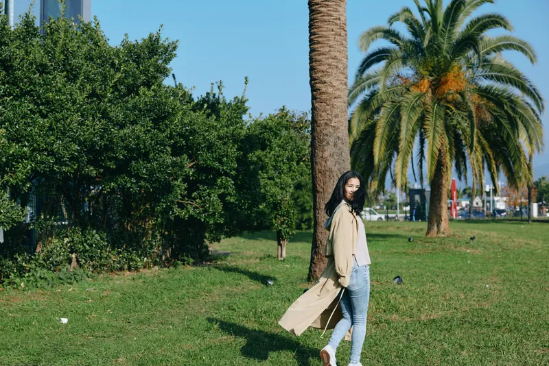 Woman walking in a sunny park with palm trees, wearing a beige trench coat and jeans, casual outdoor scene, relaxed mood, greenery and blue sky, candid lifestyle moment, soft breeze and warm light.