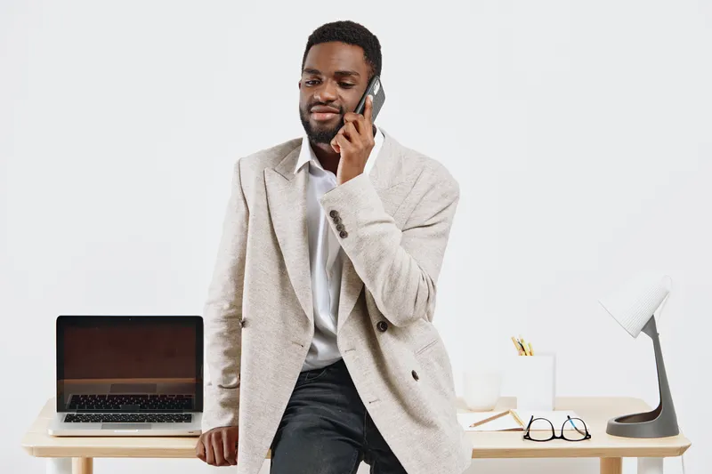 Confident man in beige blazer and white shirt smiling while talking on mobile phone in modern office setting with laptop, glasses, and desk accessories on white background. Business and communication 