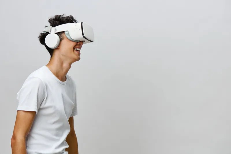 Молодой человек, погружённый в виртуальную реальность, в VR-шлеме и наушниках, улыбается от волнения на фоне минималистичного светло-серого фона, представляя будущее интерактивных развлечений и…