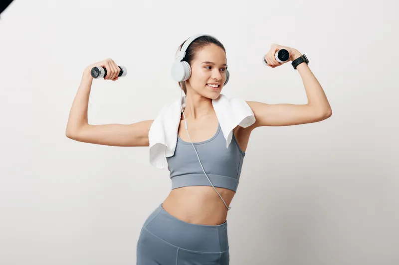 Woman in grey athletic set poses with dumbbells, headphones on and a towel over her shoulders, flexing arms in a bright studio for fitness and sport training.