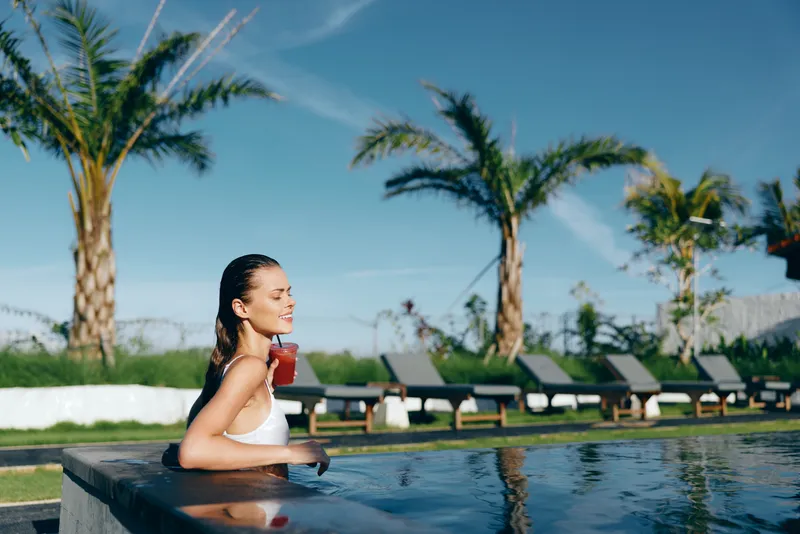 Bright poolside scene with a woman relaxing by the water, sipping a red drink, sunlit palm trees in the background, casual summer vibe, peaceful leisure lifestyle moment
