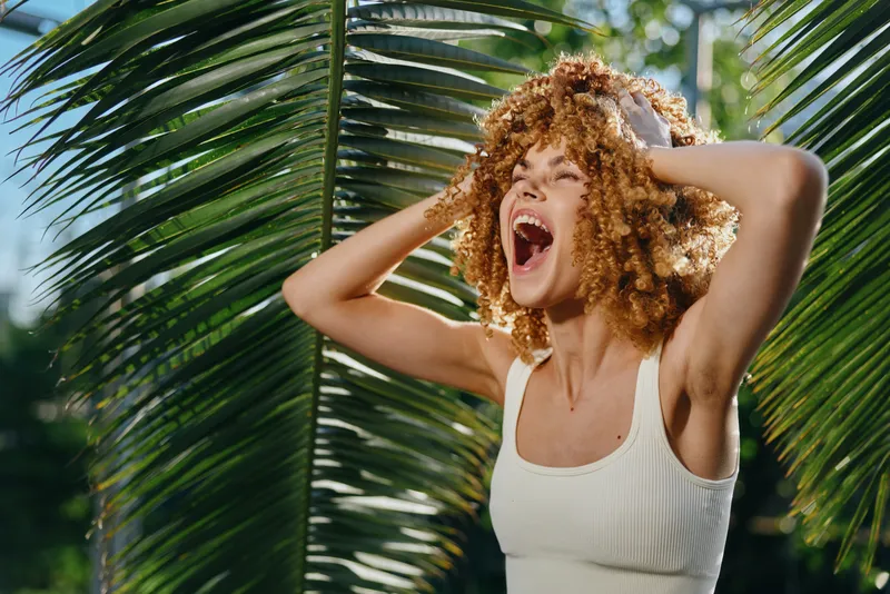 Excited woman with curly hair shouting joyfully outdoors, surrounded by tropical palm leaves and bright natural sunlight, wearing a white sleeveless top.
