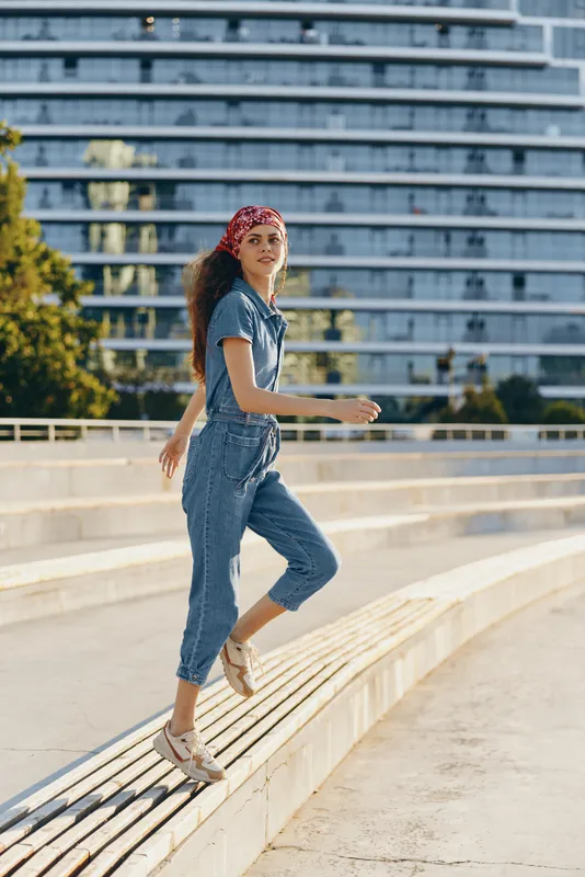 lifestyle woman in modern boho-western denim street style wearing casual outfit and red bandana, posing outdoors near urban architecture in film color tones