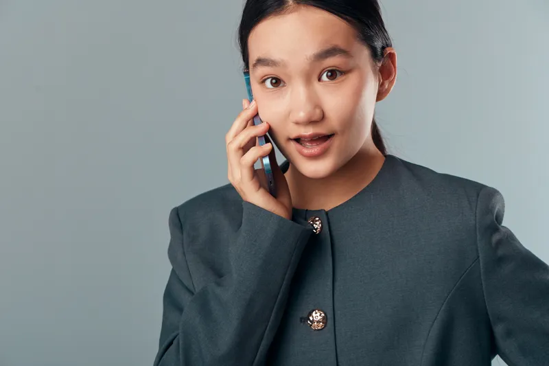Young woman talking on mobile phone with surprised expression wearing gray blazer isolated on gray background. Communication, technology, emotion, portrait, connection and casual style concept.