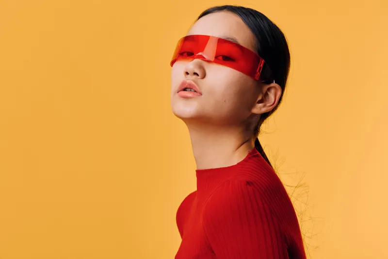 Portrait of woman wearing futuristic red sunglasses and red sweater against orange background. Fashion style, modern look with minimal makeup and sleek hair tied back, studio shot.