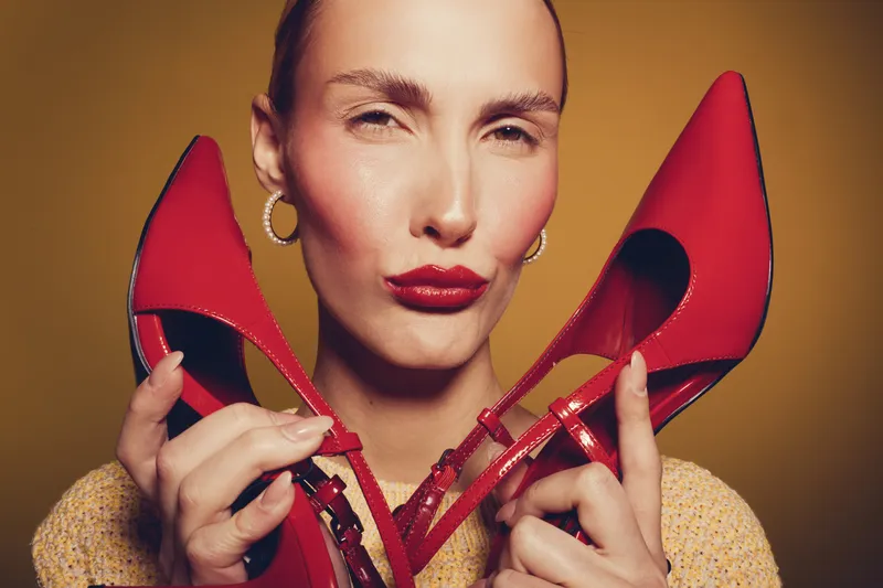 Close-up of confident woman holding red high heels near face against yellow background. Fashion concept with bold makeup and stylish accessories in studio lighting.
