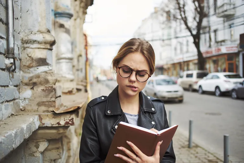 Счастливая женщина в кожаной куртке с книгой в руке идет по улице в городе