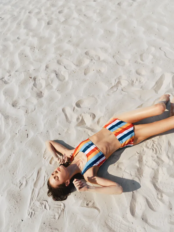 Sunlit figure lying on white sand wearing a striped swimsuit, relaxed pose, warm tones, soft shadows, and a carefree summer mood against bright light and airy sky above.