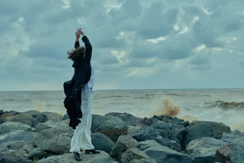 Couple dancing on a rocky shoreline by the sea, wind lifting their clothing, capturing a romantic, dynamic moment against a moody cloudy sky.