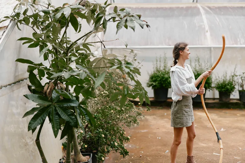 Young woman in a cozy greenhouse wearing a white hoodie and olive skirt, engaged in gardening with a curved wooden tool, surrounded by greenery
