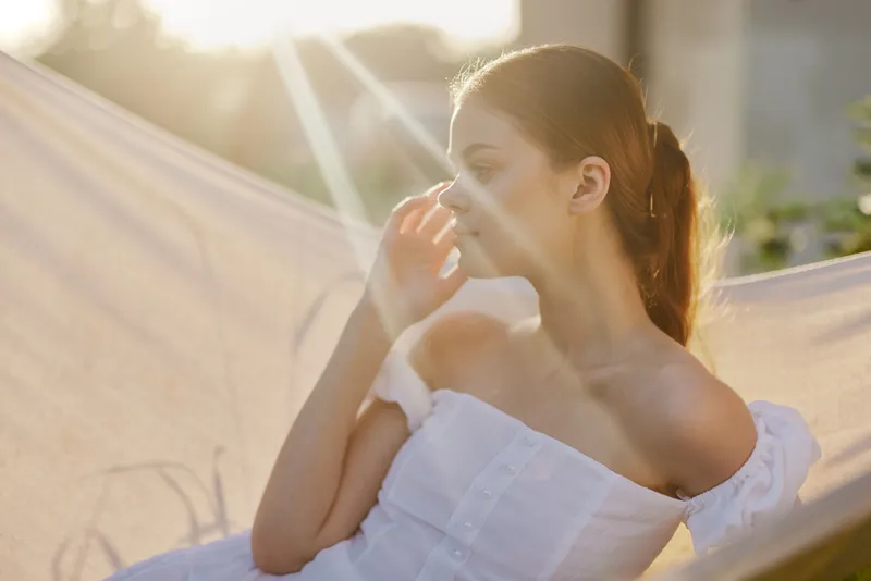 A contemplative young woman in a white off shoulder dress, enjoying a serene moment in a sunlit hammock, surrounded by greenery during golden hour