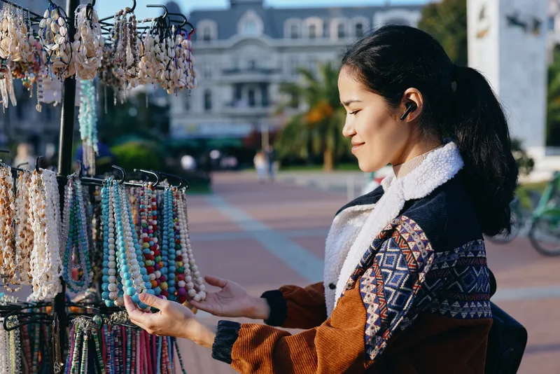 Woman browses colorful bead jewelry at an outdoor market stall, selecting bracelets and necklaces, urban plaza behind, casual knitwear, vibrant textures and artisanal craft display