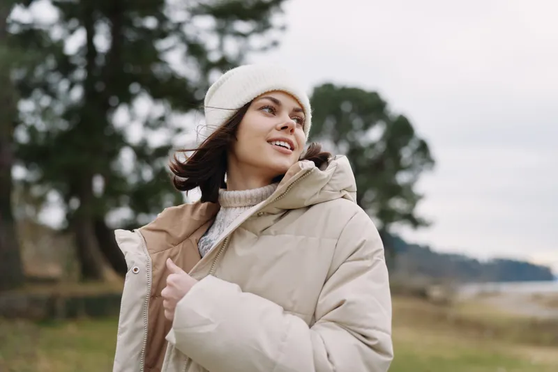 Woman in a beige puffer coat and white beanie smiles while looking up in an open field, with trees and a cloudy sky in the background.