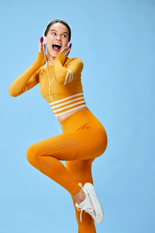 Female athlete in orange athletic wear jumping with joy against a bright blue background, headphones in, showcasing fitness energy and sporty style. The dynamic pose conveys movement, confidence, and