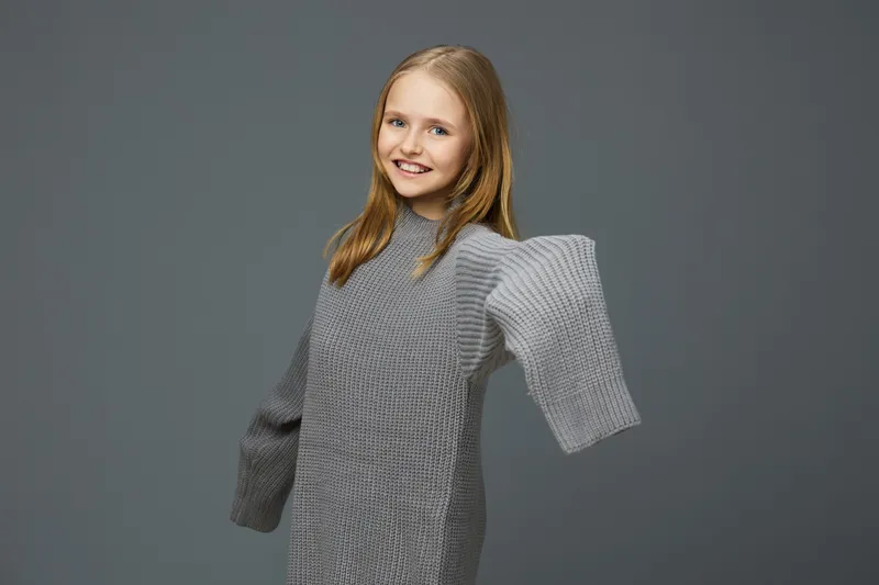 A cheerful young girl in a cozy gray oversized sweater poses against a muted gray background, capturing a moment of joy and comfort in a minimalist style