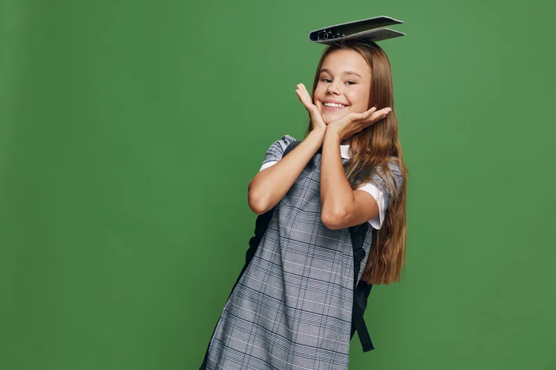 Schoolgirl smiling with hands near face and notebook on head wearing backpack standing in front of green background. Child with long hair and uniform for education concept.