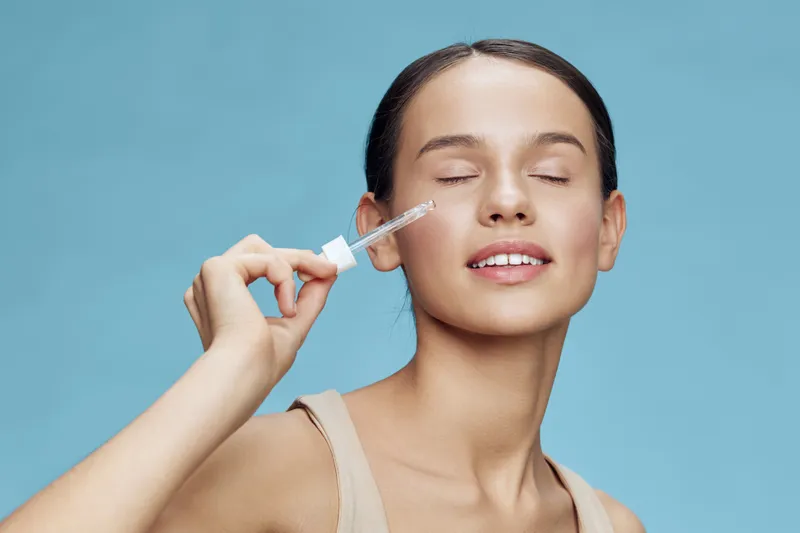Young woman applying fresh serum on her face, smiling with closed eyes against a light blue background, emphasizing skincare and natural beauty