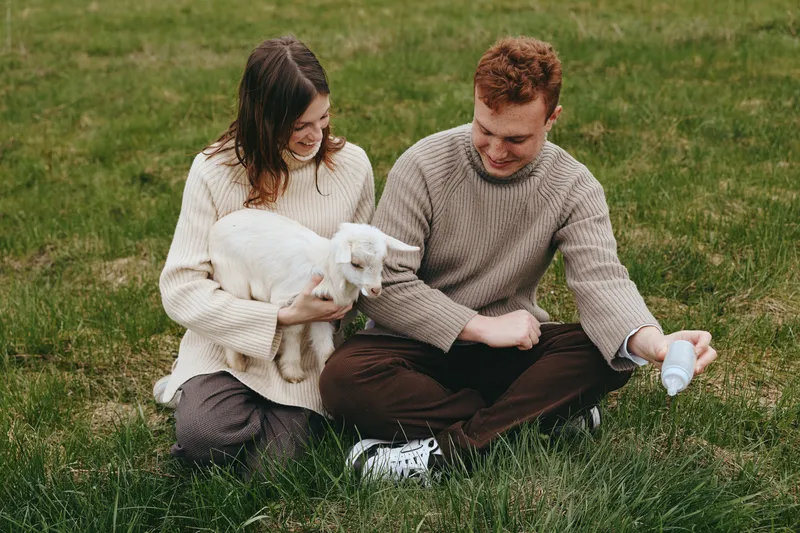 A couple sit on the grass outdoors, cuddling a white goat kid while a bottle feeds in a serene rural moment. A gentle, affectionate scene highlighting lifestyle, animal care, and family bonding in