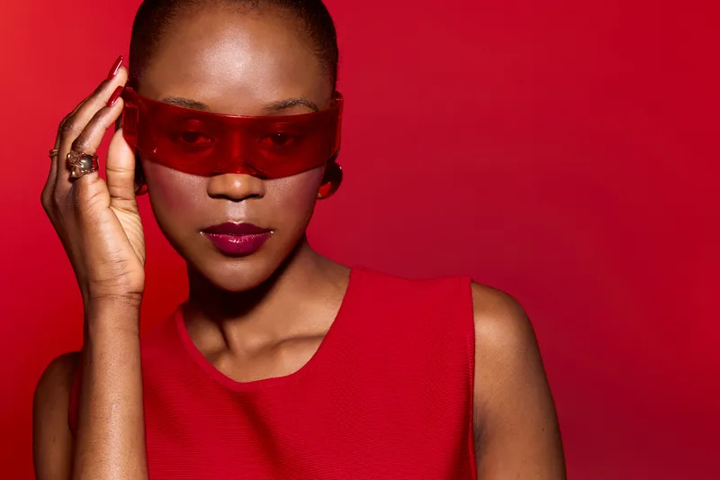 Closeup of a confident black woman with dark skin wearing red sunglasses and a red sleeveless top on a red background. Fashion concept with bold colors and modern style.