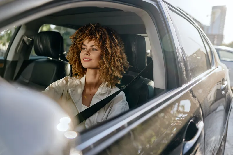 Young woman driving car safely with seatbelt on, relaxed expression, natural light, curly hair, urban background, daylight.