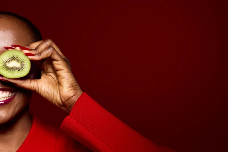 Smiling black woman with dark skin holds kiwi slice near eye on red background. Concept of healthy lifestyle, fruit, nutrition, and natural beauty with vibrant color contrast and happiness.