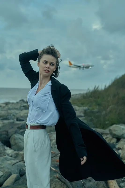 Woman on a rocky coastline, wind catching her coat, ocean horizon behind and distant airplane in the sky, stylish outdoor moment