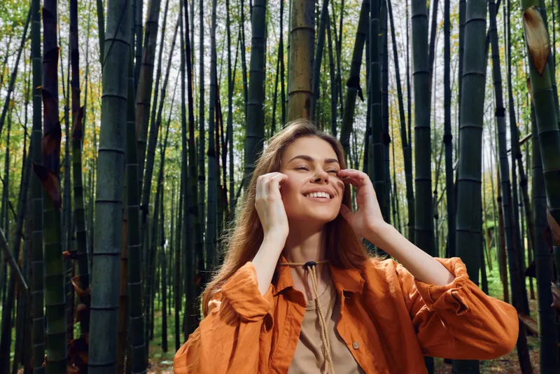 Woman in a bamboo forest smiling with eyes closed, enjoying nature and relaxation. Casual model in orange jacket breathes fresh air among tall green trees in a serene grove.
