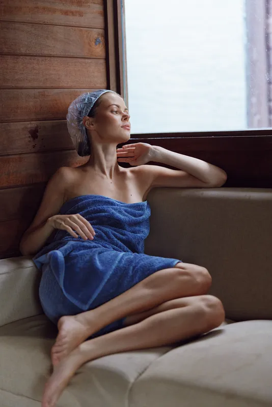 relaxed woman in blue towel sitting by a window, enjoying natural light in a cozy wooden room, calm mood, wellness and self-care atmosphere.