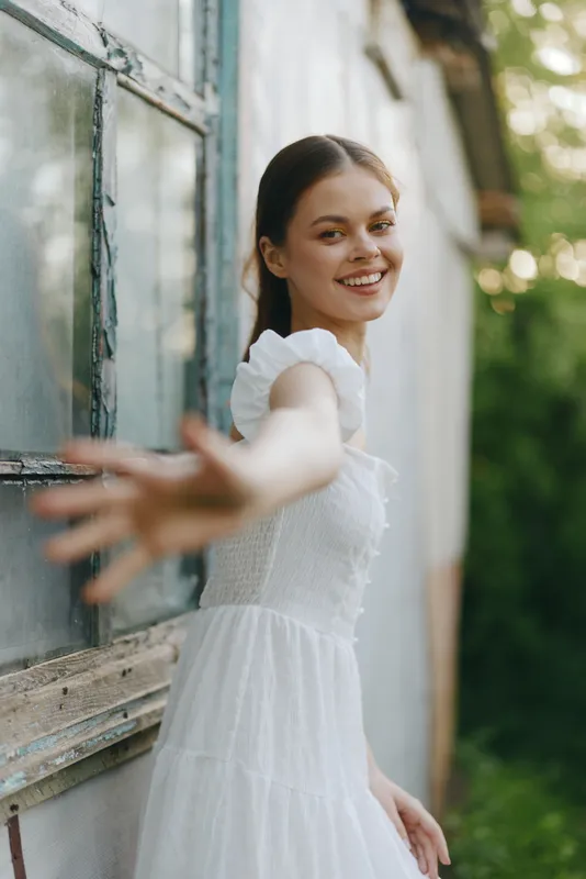 Beautiful young woman in white dress reaching out in a green outdoor setting, expressing joy and positivity while surrounded by nature