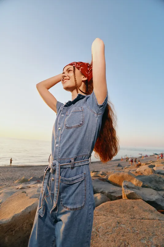 smiling woman in denim jumpsuit with red bandana enjoying sunset by rocky beach with ocean and clear sky in warm light