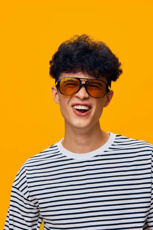 young man wearing oversized sunglasses and striped shirt smiling against a vibrant orange backdrop, casual and playful mood, modern fashion style
