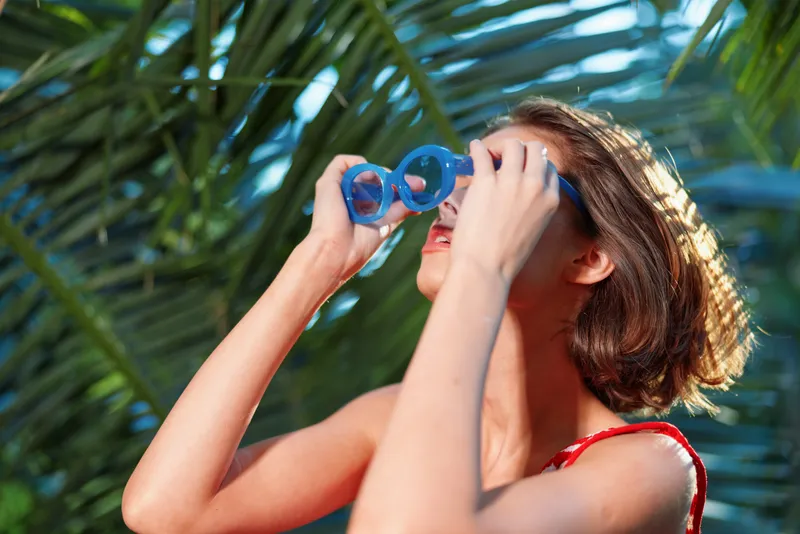 A cheerful woman uses binoculars in a sunny outdoor setting. She wears a red swimsuit top, sunlit skin, and lush tropical leaves surround her, creating a vibrant, adventurous, and playful mood.