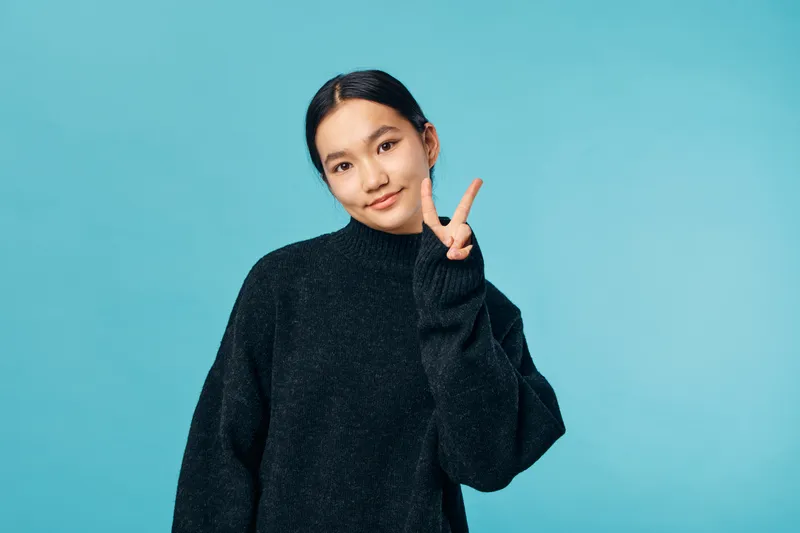 Young woman posing with peace sign wearing black sweater against blue background, smiling gently, portrait showing confident expression with casual style and neat hair.
