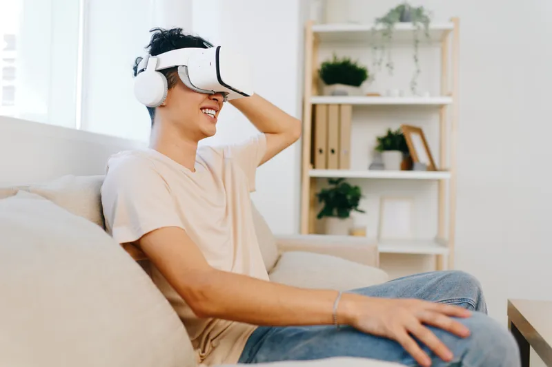 Молодой человек в VR-гарнитуре, улыбающийся и погружённый в виртуальный опыт, сидит на бежевом диване в светлой, просторной гостиной с растениями и полками на заднем плане