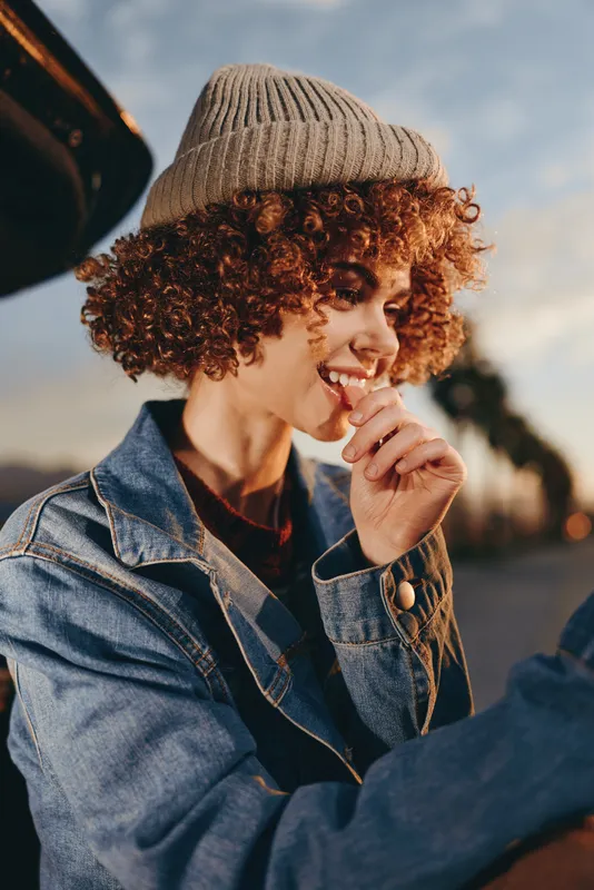 Woman lifestyle outdoors, smiling woman, curly hair, knit hat, denim jacket, golden sunset light, relaxed pose, hand near lips, warm casual mood, park path bokeh, gentle breeze