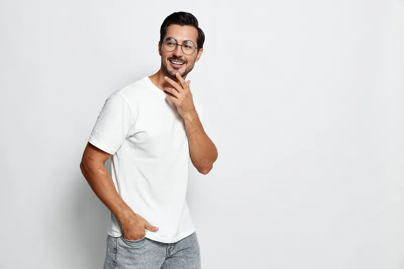 Smiling cheerful young man wearing white t-shirt and glasses poses playfully while looking at the camera, showcasing a confident and relaxed demeanor against a plain background.