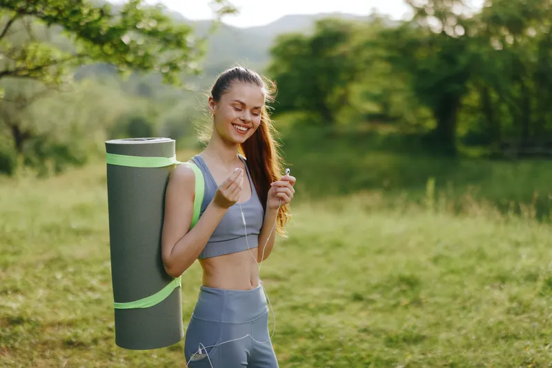 Young woman with yoga mat enjoying nature, smiling in yoga outfit, green background, promoting healthy lifestyle, fitness and well being